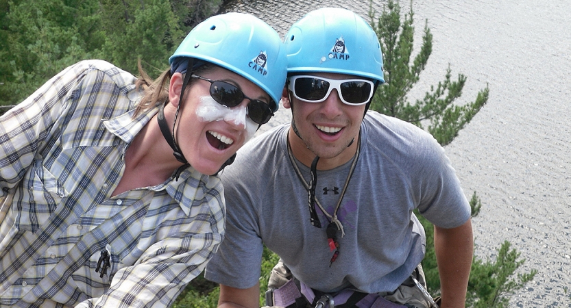 Two people wearing helmets smile at the camera. They appear to be at a high elevation. There is water below them.